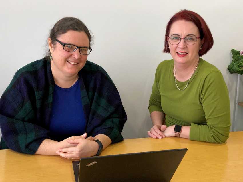 Heather and Deborah are sitting at a desk looking at the camera.  A laptop faces them in the foreground.