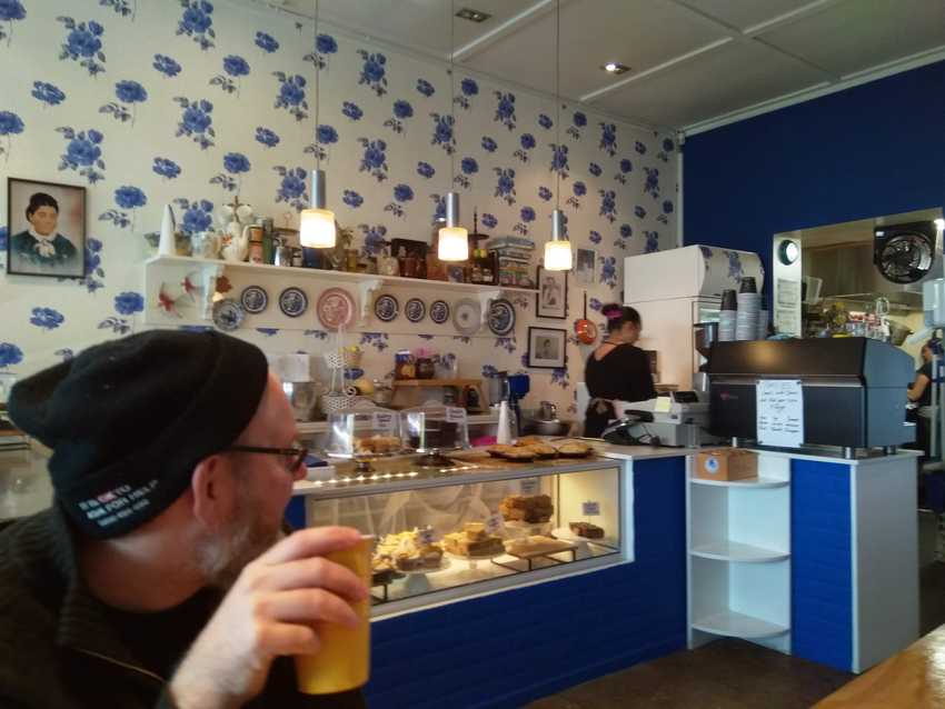 inside Blue Rose Cafe.  Wallpaper behind the counter is white with blue roses.  In the foreground a person sips coffee looking away from the camera.