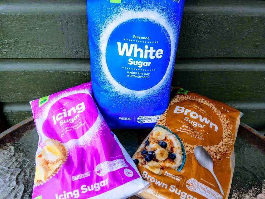 Three bags of sugar. A blue bag of white sugar leaning against a green wall. A pink bag of icing sugar and a brown bag of brown sugar lying along the flat surface.